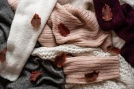 Ovanti Strickdesign – Background with warm sweaters. Pile of knitted clothes with autumn leaves, warm background, knitwear, space for text, Autumn winter concept.