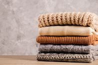Ovanti Strickdesign – Bunch of knitted warm pastel color sweaters with different knitting patterns folded in stack on brown wooden table, grunged texture wall background. Fall winter season knitwear.