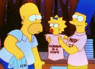 Die Simpsons – Stolz zeigt Marge (re.) ihrem Mann Homer die T-Shirts, die sie gekauft hat. Darauf steht: "Homer ist ein Dummkopf!"