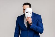 Reingelegt! – Bearded man holding white mask, covering face, standing with serious expression, multiple personality disorder, wearing official style suit.