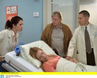 In aller Freundschaft – Dr. Kaminski (Udo Schenk, rechts) und Burkard Dreher (Arved Birnbaum, 2. von rechts) sind auf dem Weg zu Iris Dreher (Nadja Engel, liegend), als diese von Dr. Eichhorn (Cheryl Shepard) aus dem Zimmer gerollt wird. Iris Dreher ist kollabiert und muss sofort notoperiert werden.