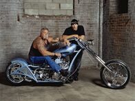 American Chopper – Paul Sr. sits on Paul Jr.'s blue auction bike while Paul Jr. leans on bike. As seen on AMERICAN CHOPPER.