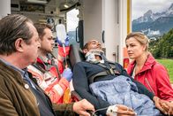 Watzmann ermittelt – Jerry Paulsen (Peter Marton, liegend) hatte einen Unfall: An seinem Dienstwagen haben die Bremsen versagt. Johanna (Ines Lutz, r.) und ihr Vater Benedikt Beissl (Andreas Geibel, l.) sind zur Stelle, wie der verletzte Jerry in den Krankenwagen geladen wird. Sanitäter (Komparse, 2.v.l.)