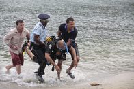 Death in Paradise – Phil Owen (Sean Gallagher, l.), Dwayne (Danny John-Jules, 2.v.l.) und Mark Lightfoot (O-T Fagbenle) tragen den leblosen Körper von Benjamin Lightfoot (Alex Lanipekum) aus dem Meer.