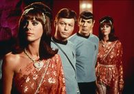 Raumschiff Enterprise – Dr. McCoy (DeForest Kelley, 2.v.l.) und Mr. Spock (Leonard Nimoy, 2.v.r.) beobachten leicht abgestoßen die fremde Welt von Harry Mudd. Der Herrscher hat sich eine Bevölkerung aus Androiden geschaffen, über die er verfügen kann. Die Roboter-Frauen (Darstellerinnen unbekannt) gleichen einander wie ein Ei dem anderen.