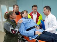 In aller Freundschaft – Paul Menke (Pierre Kiwitt, liegend) ist plötzlich mit starken Magenkrämpfen in seinem Fischgeschäft zusammengebrochen. Die Notärzte (Komparsen) bringen ihn in die Sachsenklinik. Maja Jäger (Ruth Moschner, links), die seinen Fischladen beliefert, lässt Paul keine Minute allein. Als Dr. Roland Heilmann (Thomas Rühmann, rechts) sich erkundigt, ob Paul vielleicht etwas Falsches gegessen haben könnte, kommt Paul ein Verdacht: Hatte Lebensmittelkontrolleur Smolka doch recht?