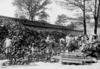Ein Tag in Auschwitz – Die Habe der Menschen, deren Transport „abgefertigt“ wurde wird im Materiallager „Kanada“ von Häftlingen sortiert. Im Hintergrund qualmt eine Grube, in der Leichen verbrannt werden.