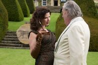 Father Brown – Eine Prostituierte von Madame Chania (Ronni Ancona, l.) wurde ermordet. Der Verdacht fällt sofort auf den Hotelgast Dr. Barashi (Michael Culkin, r.), der im Hotelgarten Schießübungen abhält.