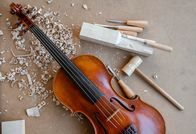 Heimatleuchten – Rotbraune Geige auf dem Schreibtisch eines Geigenbauers. Schnitzwerkzeuge, Sägemehl und Holzhammer.