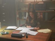 Love Kills – Melissa Bayern is (Margaret) sitting at her desk reading a book.