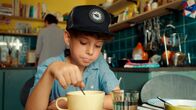 Geschichten vom Franz – Ein Junge mit Basecap sitzt an einem Küchentisch und isst Müsli mit Milch aus einer großen Tasse.
