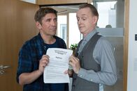 Löwenzahn – Äußerst skeptisch und mit spitzen Fingern überreicht Amtmann Kluthe (Holger Handtke) das Regelwerk für den Ideenwettbewerb "Bärstadt bekommt eine neue Schule" an Fritz (Guido Hammesfahr).