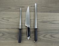 We love Cooking – Foto eines einzelnen großen Küchenmessers und zweier Schärfgeräte auf gealtertem Weißeschenholz