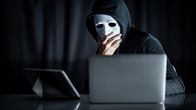 Reingelegt! – Mystery male hoodie hacker holding white mask looking at laptop computer. Anonymous social masking. Ransomware cyber attack or internet security concepts