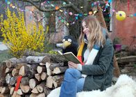 Rabe Rudi - Vorlese-Geschichten – Milena Tscharntke und Rabe Rudi sitzen auf einem Holzstapel vor einem Haus und lesen gemeinsam in einem Buch.