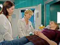 In aller Freundschaft – Tanja Wassmuts (Victoria Deutschmann, rechts) Leber reagiert sehr empfindlich auf die im Fotolabor verwendeten Chemikalien. Sie war deshalb schon oft in der Klinik. Dr. Eichhorn (Cheryl Shepard, links) und Dr. Globisch (Andrea Kathrin Loewig) versuchen ihr zu erklären, dass sie mit ihrem Job ihr Leben aufs Spiel setzt.