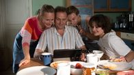 Für immer Eltern – Erwachsene Familienidylle: Anja Wagner (Anja Schneider, l.), ihr Mann Michael (Devid Striesow, 2.v.l.) und ihr Sohn Niklas (Max Schimmelpfennig, 2.v.r.) sprechen per Bildschirm am Frühstückstisch mit Niklas' Schwester Stella. Niklas' Freundin Alina (Anouk Elias, r.) verfolgt das Gespräch amüsiert.
