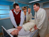 In aller Freundschaft – Als Hans-Peter Brenner (Michael Trischan, links) seiner Mutter sagt, dass er die Stelle in der Klinik wieder gekündigt hat, um sich um sie zu kümmern, kommt es zum Streit und Luises (Monika Lennartz, liegend) Zustand eskaliert. Brenner schickt Schwester Yvonne (Maren Gilzer, 2. von links) und Oberschwester Ingrid (Jutta Kammann, 3. von links) los, um Medikamente und den Reanimationswagen zu holen. Selbst als Dr. Heilmann (Thomas Rühmann) im Krankenzimmer eintrifft, lässt ihn Brenner nicht an seine Mutter ran, bis sie wieder einen Sinusrhythmus hat.