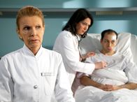 In aller Freundschaft – Oberschwester Ingrid (Jutta Kammann, vorn) hat Benjamin Helbig (Tom Lass, rechts) versprochen, dass der Professor seinem jahrelangen Leiden ein Ende machen kann und einen neuen Therapieansatz für die Behandlung seiner Rheumatoiden Arthritis finden wird. Doch dem war nicht so. Benjamin beginnt aufzugeben, noch dazu wurde ihm nun auch noch eine Herzbeutelentzündung diagnostiziert. Als Benjamin wieder Schmerzen hat und Dr. Elena Eichhorn (Cheryl Shepard) ihn untersucht, stellt sie fest, dass sich Benjamins Entzündung verschlimmert hat. Er muss sofort in den Schockraum.