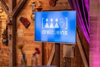 dreizueins – Logo von "dreizueins" auf einem Bildschirm in der Studiokulisse.