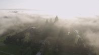 Wunderwerke der Weltgeschichte – Luftaufnahme eines großen, antiken Tempelkomplexes, teilweise von Nebel umhüllt. Mehrere Türme mit spitzen Spitzen ragen aus der üppigen Vegetation hervor. Sonnenstrahlen durchbrechen den Nebel und erzeugen eine dramatische, fast mystische Atmosphäre. Die Szene wirkt wie in den frühen Morgenstunden aufgenommen und zeigt die majestätische Architektur von Angkor Wat in ihrer natürlichen Umgebung.