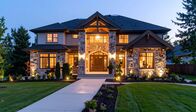 Die Kalteneggers – A luxurious twostory house, illuminated at dusk, features stone facade, large windows, landscaped lawn, and a paved walkway leading to the grand entrance.