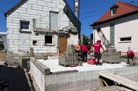 Handwerkskunst! – Maurermeister Erik Zedler und Mitarbeiter bei Hausbau. An einen bestehenden Altbau, kommt ein Neubau – gerade wird die erste Schicht Bimssteine in Mörtel gesetzt.