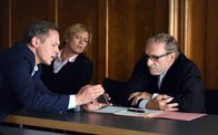 Über Land – Franz Kurz (Johannes Zirner, l.) zeigt Max Althammer (Franz Xaver Kroetz, r.) und der Protokollführerin (Michaela Heigenhauser, M.) Bilder der Zerstörung seiner Wohnung.