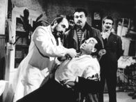 Don Camillos Rückkehr – Mit Genuss beobachtet Peppone (Gino Cervi, Mitte), wie sich sein Lieblingsfeind Don Camillo (Fernandel, sitzend) versucht, sich dem Zahnarzt zu widersetzen.