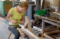 Handwerkskunst! – Ingrid Ertel sitzt auf dem Schneidstuhl ihres Großvaters und hobelt mit dem Ziehmesser Späne von der späteren Stütze für die Obstleiter.