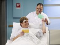 In aller Freundschaft – Hans-Peter Brenner (Michael Trischan, hinten) steht vor einer wichtigen Prüfung für sein Medizinstudium und findet keine Zeit zum Lernen. Patient Hanzer (Frank Schöbel, liegend) nimmt sich ein Herz und fragt Hans-Peter ab. Als der Patient allerdings in den OP muss, verschwinden mit ihm auch Brenners Lernkarten.